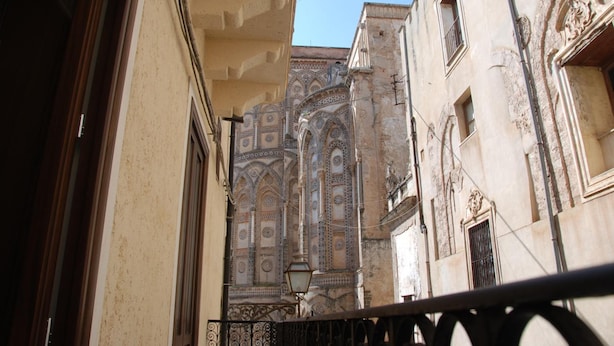 A narrow alleyway with ornate architecture, including a building with detailed tile work and a balcony with a wrought iron railing. - Locanda Re Ruggero (Monreale)