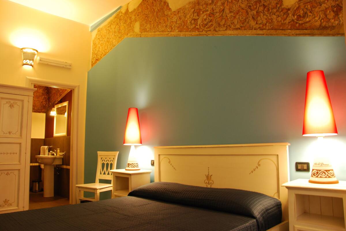 A bedroom with a bed, two bedside lamps, a nightstand, a dresser, and a bathroom visible in the background.