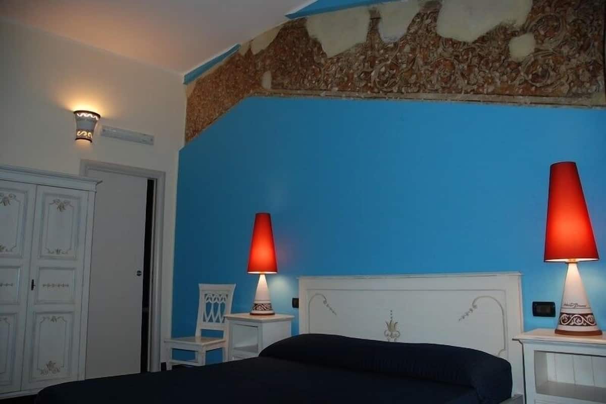 A bedroom with a blue wall, a bed with a dark cover, two bedside lamps with red shades, a white door, and a small white nightstand with a decorative item.