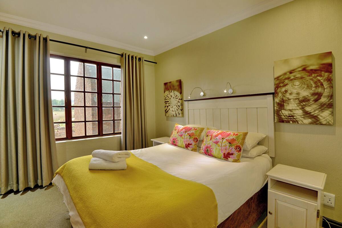 A hotel room with a bed, a nightstand, a window with curtains, and a wall with a painting.