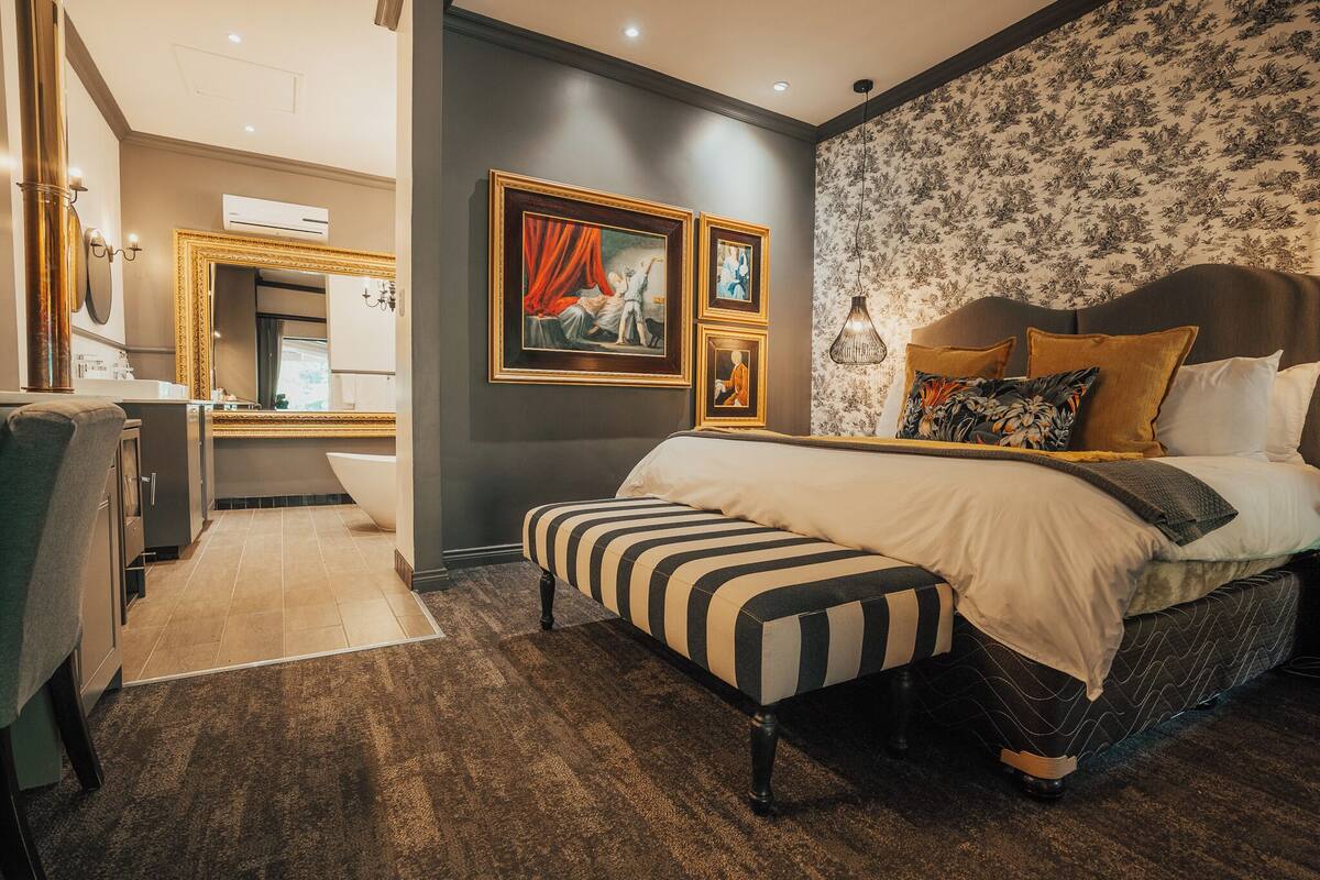 A bedroom with a large bed, a bench, a vanity with a mirror, and framed artwork on the wall.