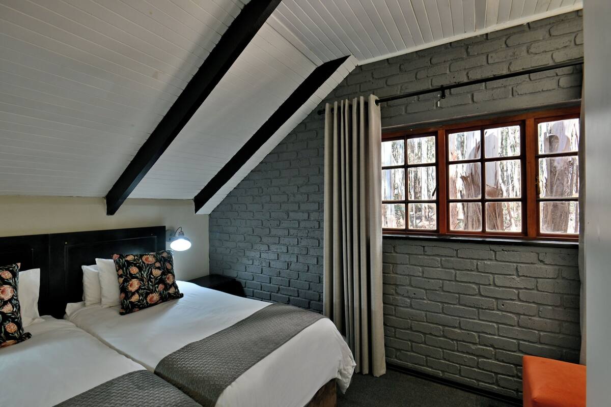 A room with two beds, a brick wall, a window with curtains, and a sloped ceiling.