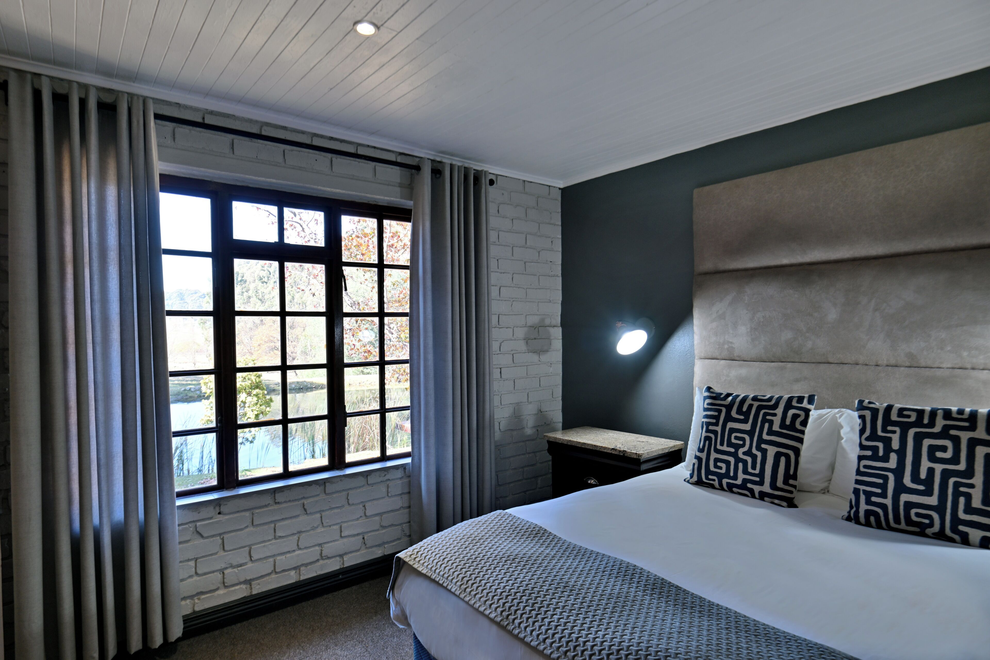 A modern hotel room with a large bed, a headboard, a nightstand, a window with curtains, and a view of the outdoors.