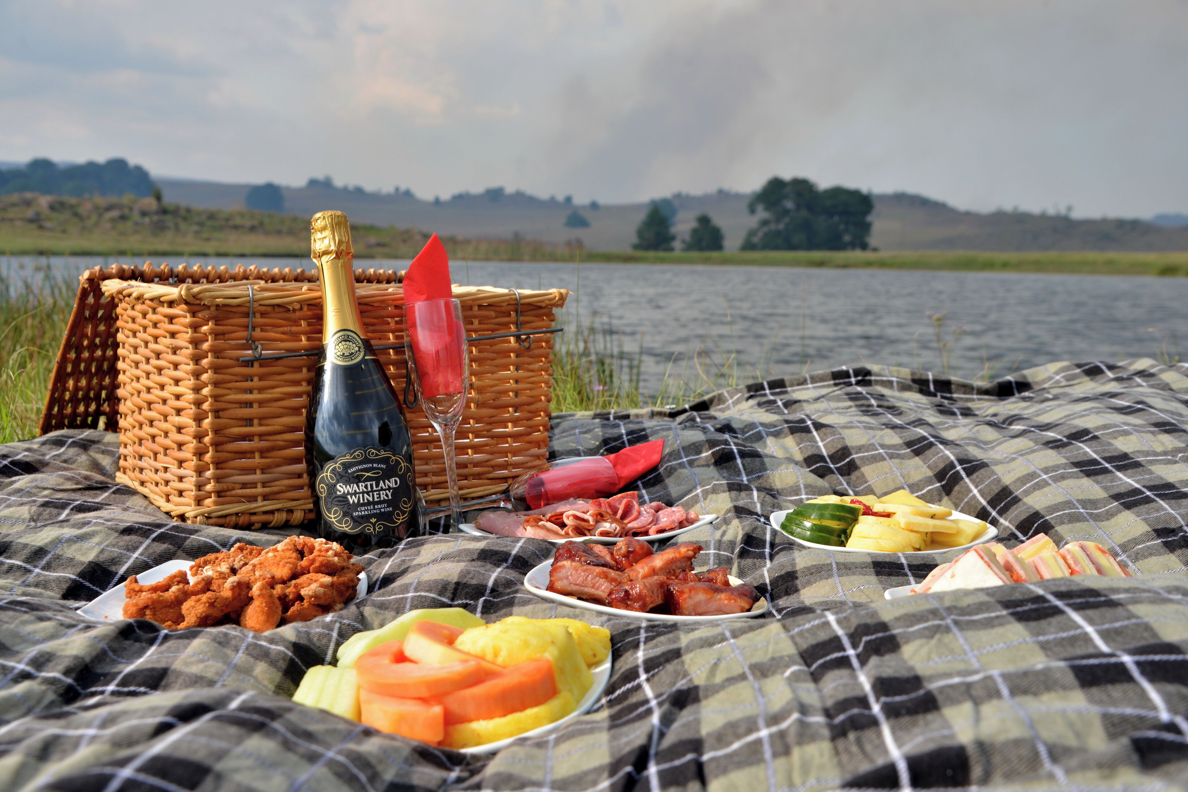 A picnic setup with a bottle of wine, a basket, and various food items on a checkered blanket by a lake.