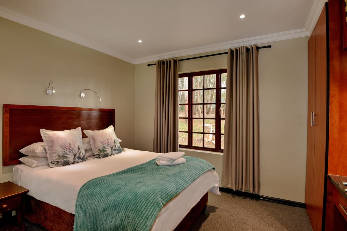A neatly made bed with a wooden headboard, a green throw blanket, and floral pillows. There is a window with curtains, a nightstand, and a wooden wardrobe.