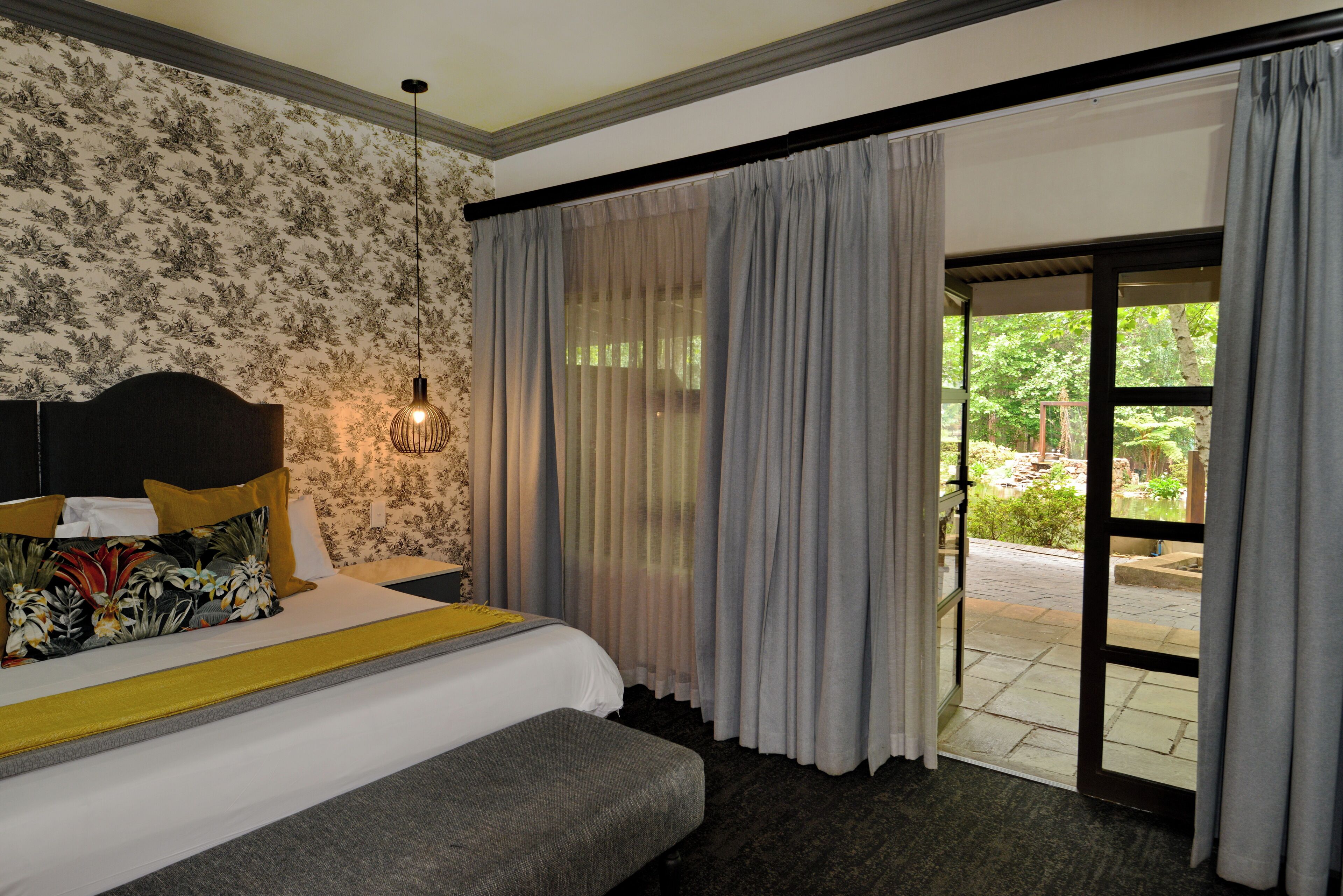 A hotel room with a bed, a bench, a nightstand, a lamp, and a view of the garden through the window.