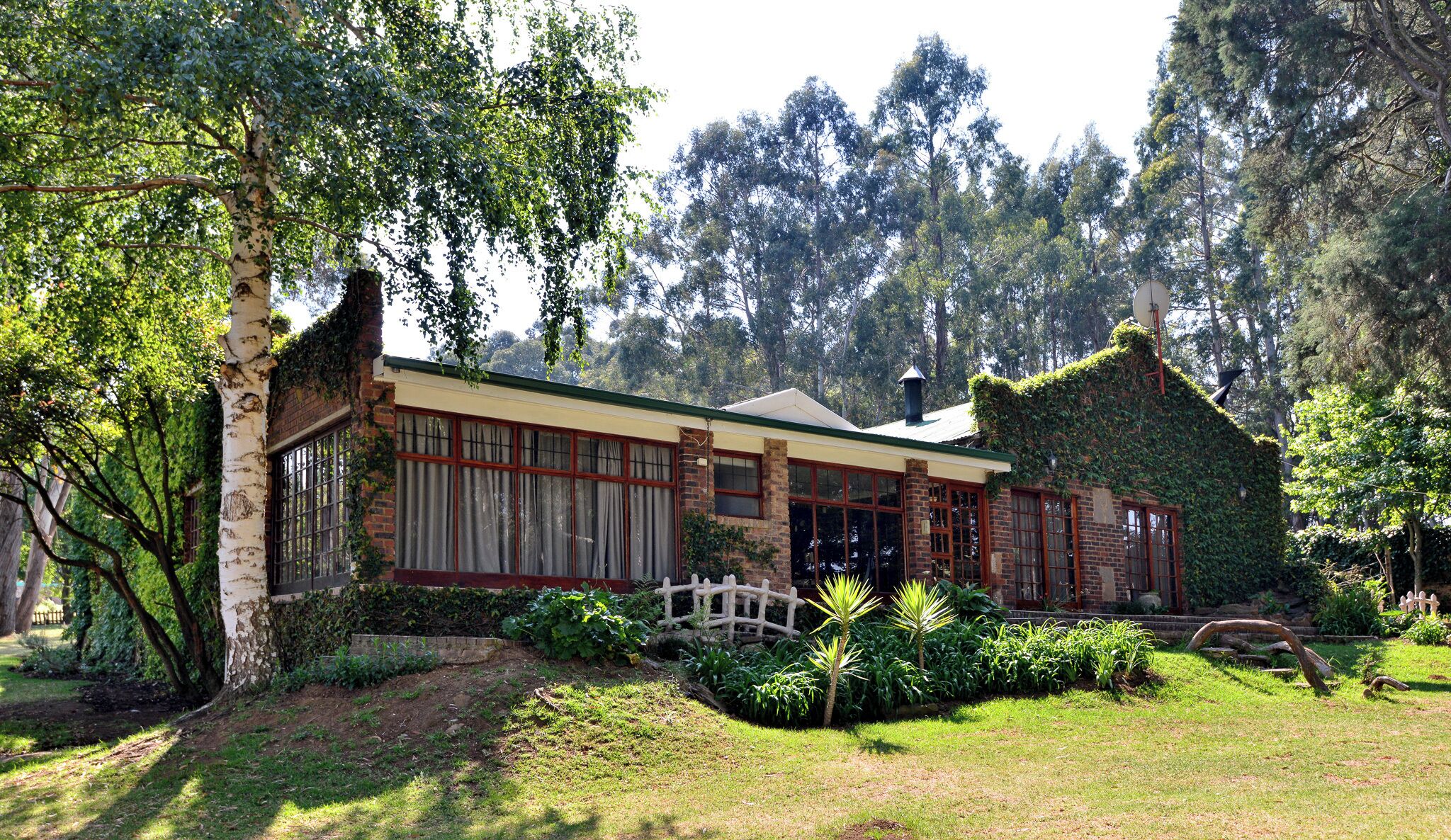 A house with a brick facade, large windows, and a garden with various plants and a small bridge.