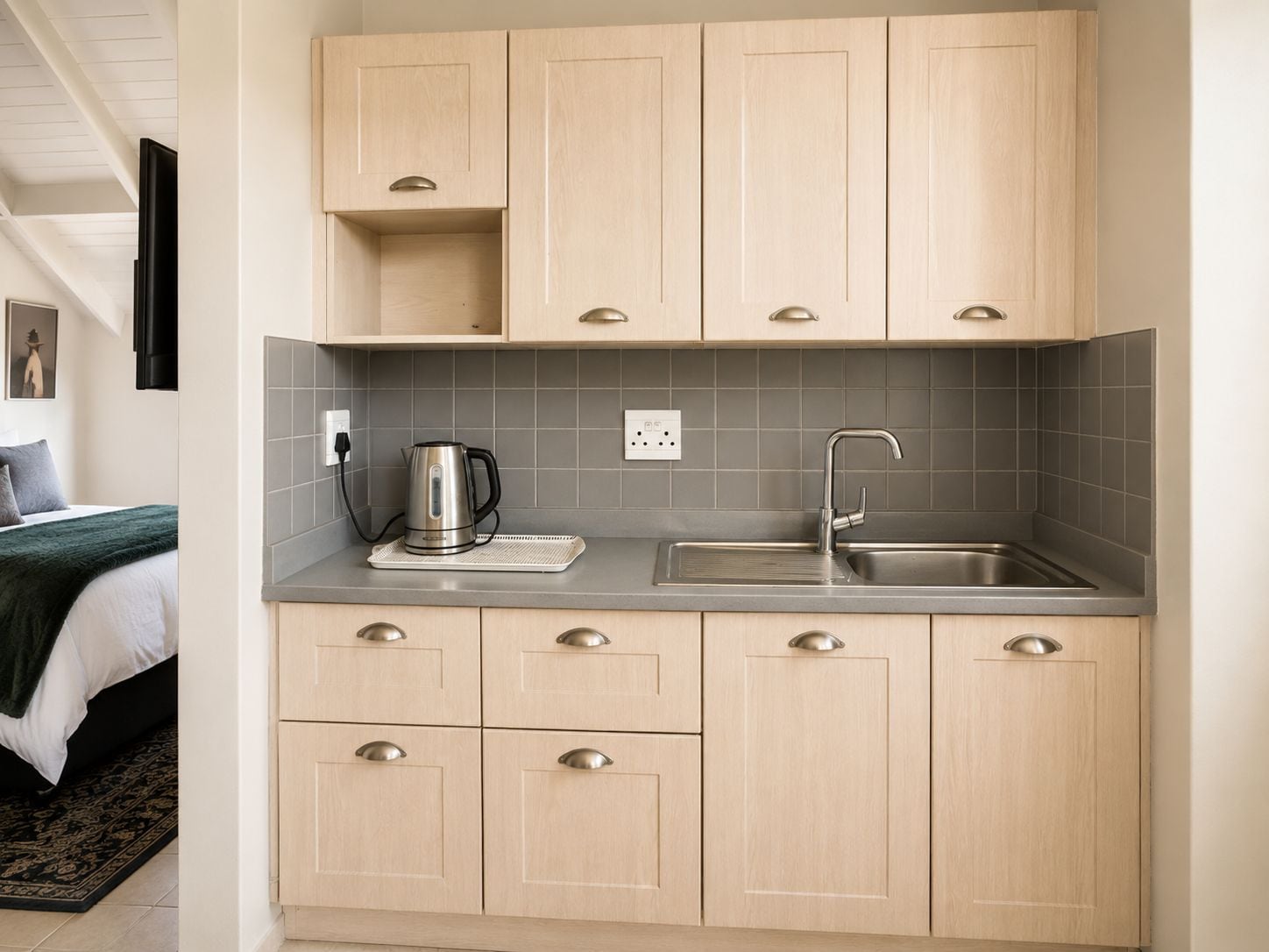A modern kitchen with light wood cabinets, a stainless steel sink, and a kettle on the countertop.