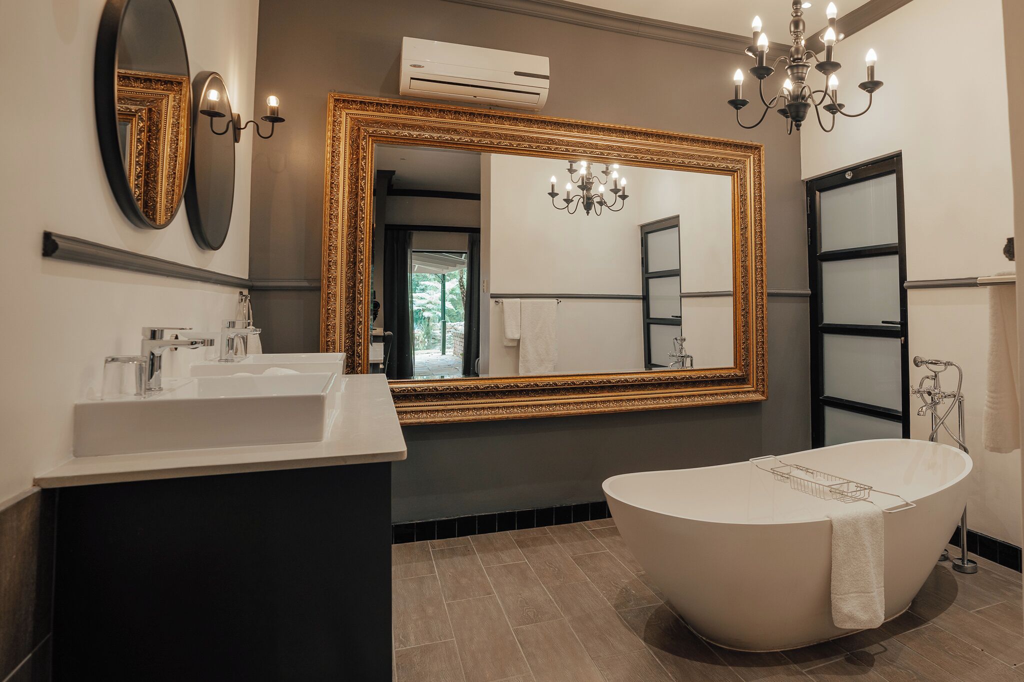 A modern bathroom with a freestanding bathtub, a large mirror with a gold frame, and a chandelier.