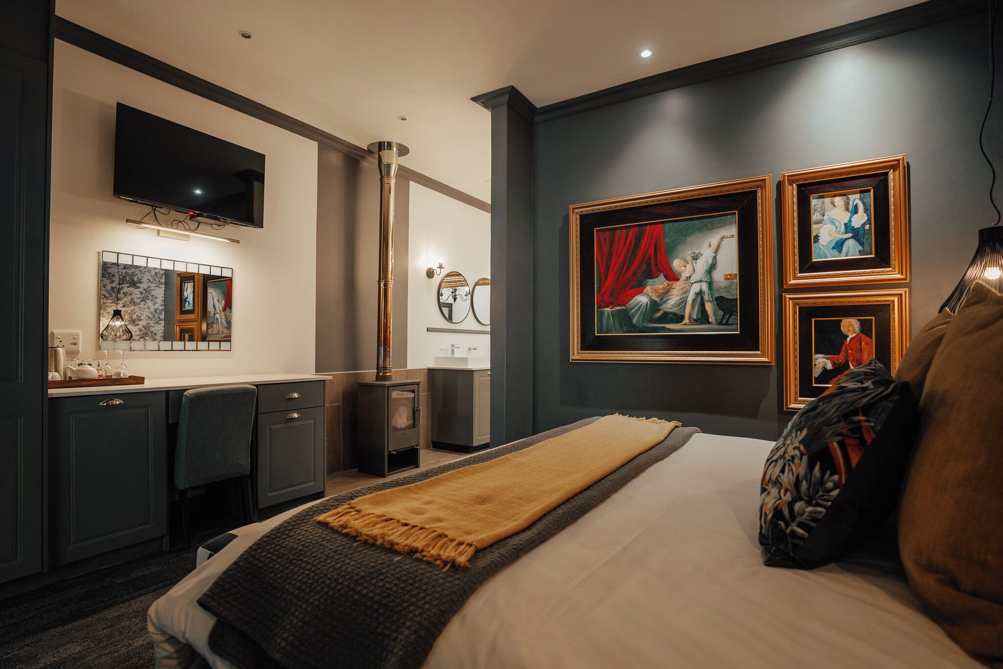 A modern hotel room with a large bed, a flat-screen TV, a kitchen area, and artwork on the walls.