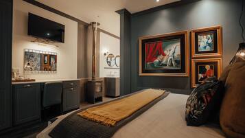 A modern hotel room with a large bed, a flat-screen TV, a kitchen area, and artwork on the walls.