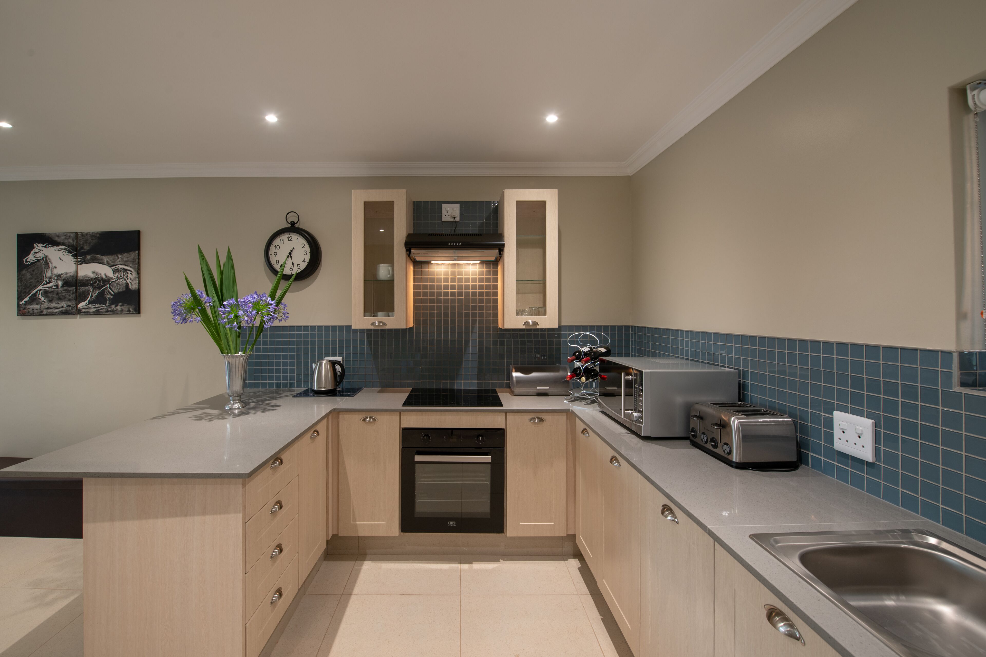 A modern kitchen with a central island, built-in oven, and a microwave. There is a clock on the wall and a vase with flowers on the island.