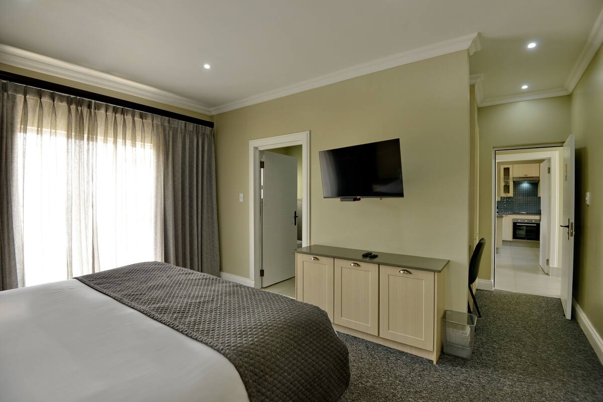 A hotel room with a large bed, a TV mounted on the wall, a small kitchenette, and a bathroom visible through an open door.