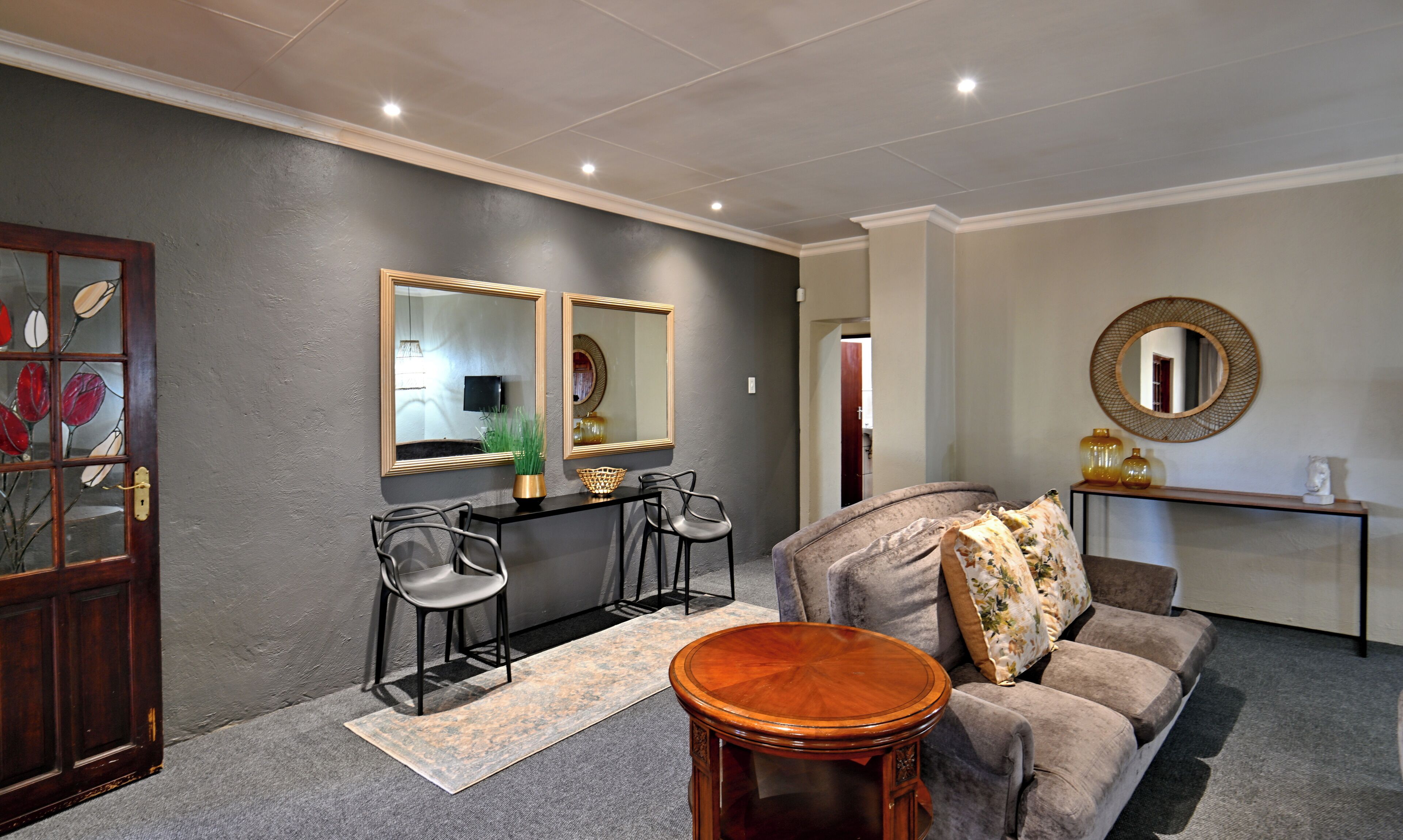 A living room with a grey wall, a wooden door, a grey sofa, a round wooden table, two black chairs, a small rug, and a mirror on the wall.