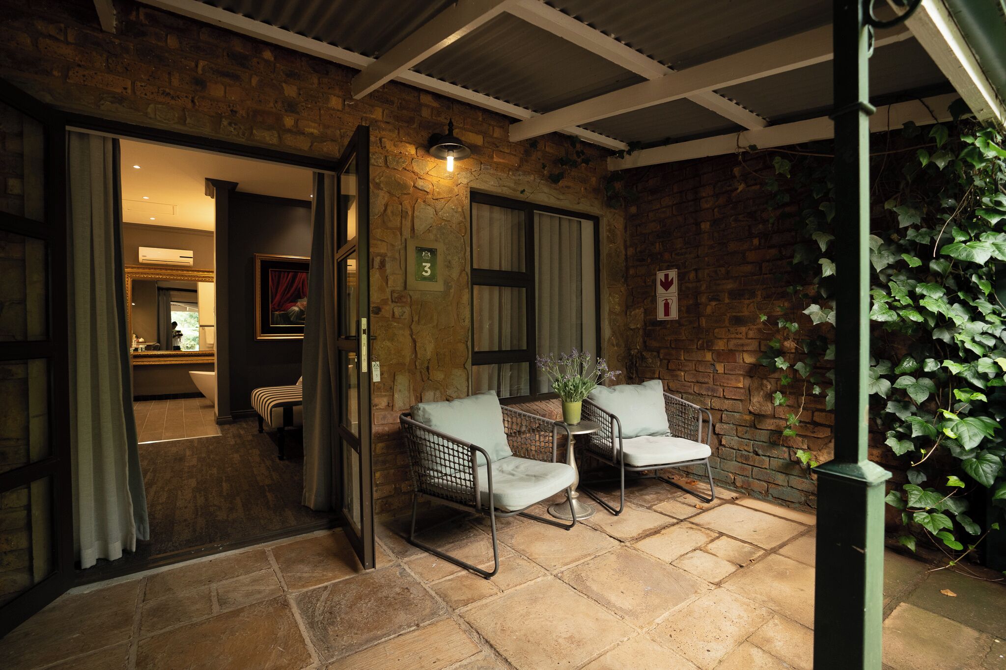 A cozy outdoor seating area with two chairs and a table, a brick wall, and a hanging plant.