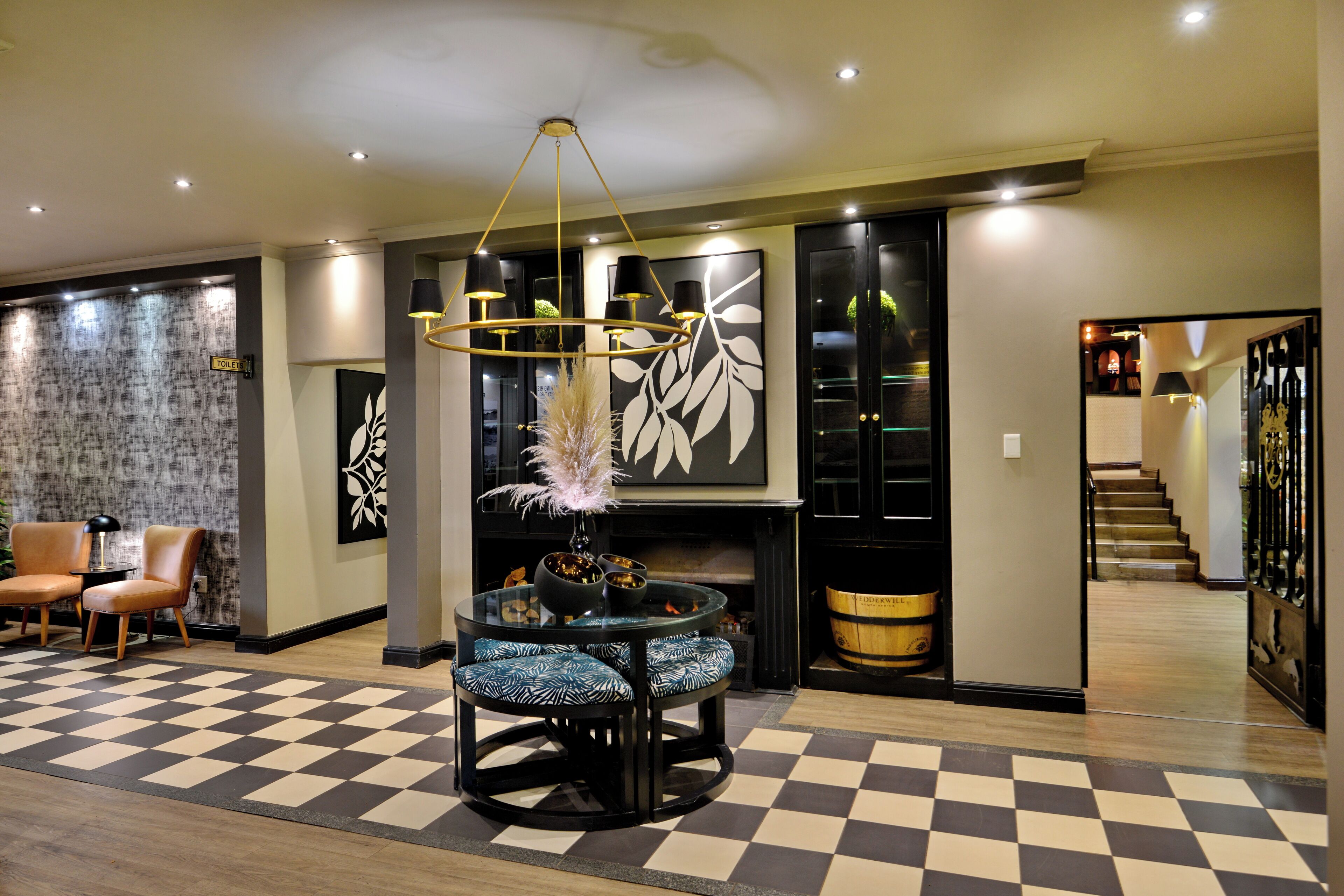 A modern lobby with a checkered floor, a round glass table with chairs, a fireplace, and a staircase in the background.
