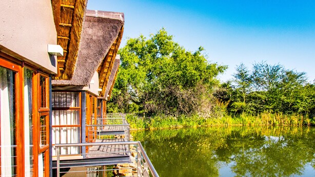 A building with a thatched roof and a wooden balcony overlooking a calm body of water. - Seasons Golf, Leisure & Spa (Hartbeespoort/ Brits)