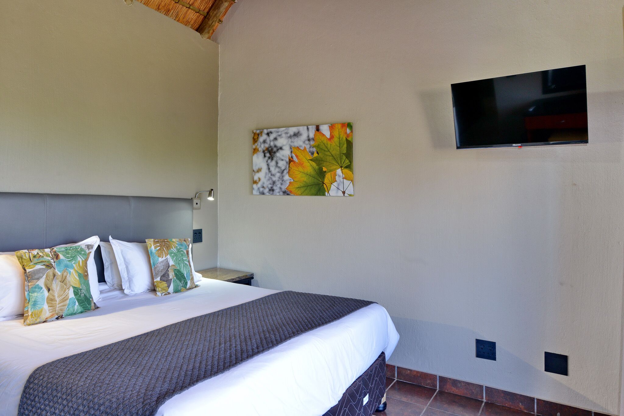 A hotel room with a bed, a flat-screen TV mounted on the wall, and a painting of leaves.