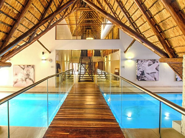 A swimming pool with a wooden deck and glass railings, flanked by walls with artwork and a staircase leading to an upper level. - Seasons Golf, Leisure & Spa (Hartbeespoort/ Brits)