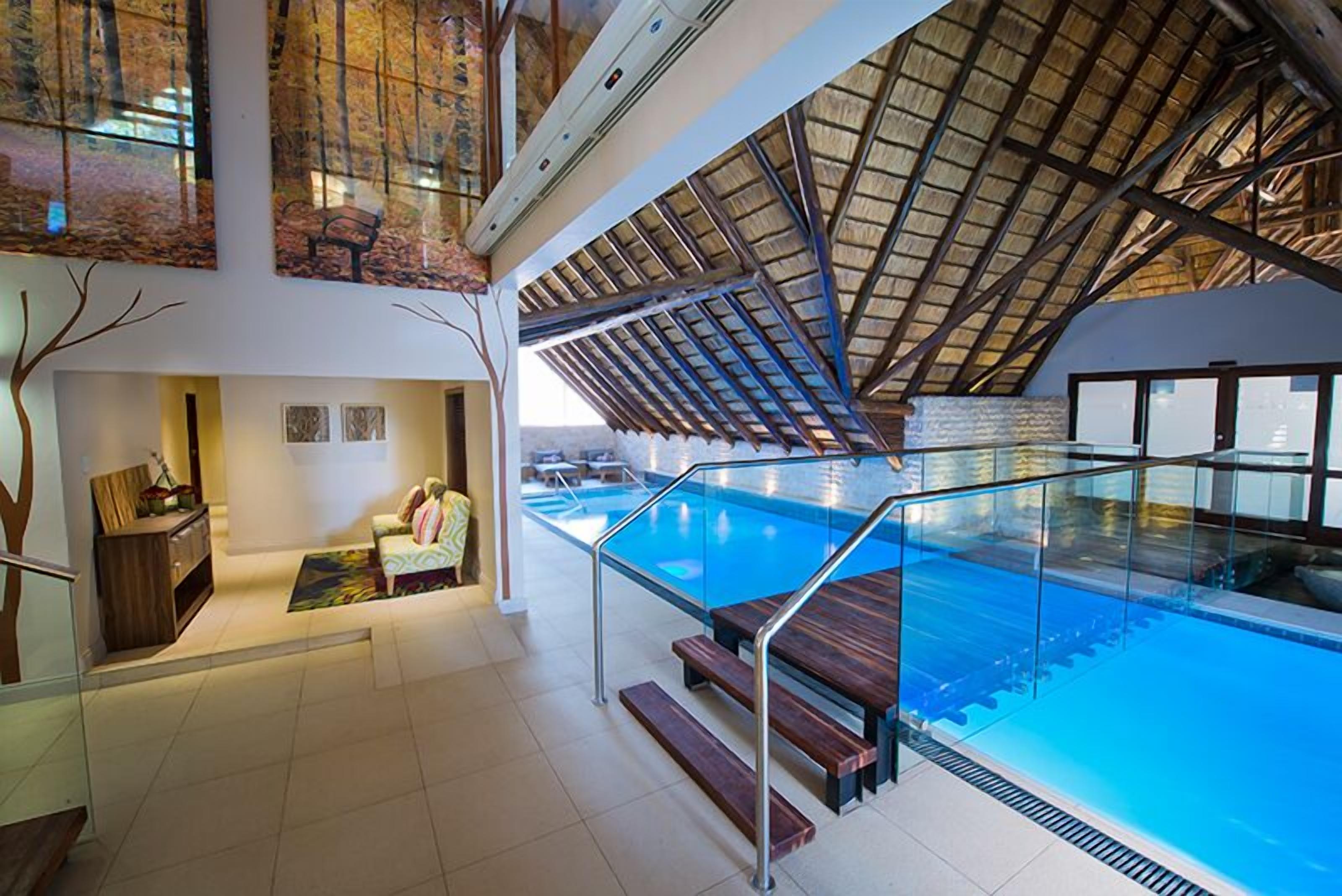 Una amplia piscina interior con barrera de vidrio, terraza de madera y techo alto con techo de paja.