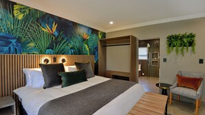 A modern hotel room with a large bed, a wooden headboard, a tropical mural, a small seating area, and a bathroom visible through a glass door. - Seasons Golf, Leisure & Spa (Hartbeespoort/ Brits)