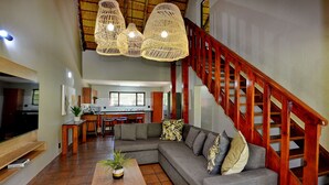 A spacious living area with a large gray sectional sofa, a wooden coffee table, and a TV on a wooden stand. A wooden staircase with a red railing leads to an upper level. - Seasons Golf, Leisure & Spa (Hartbeespoort/ Brits)