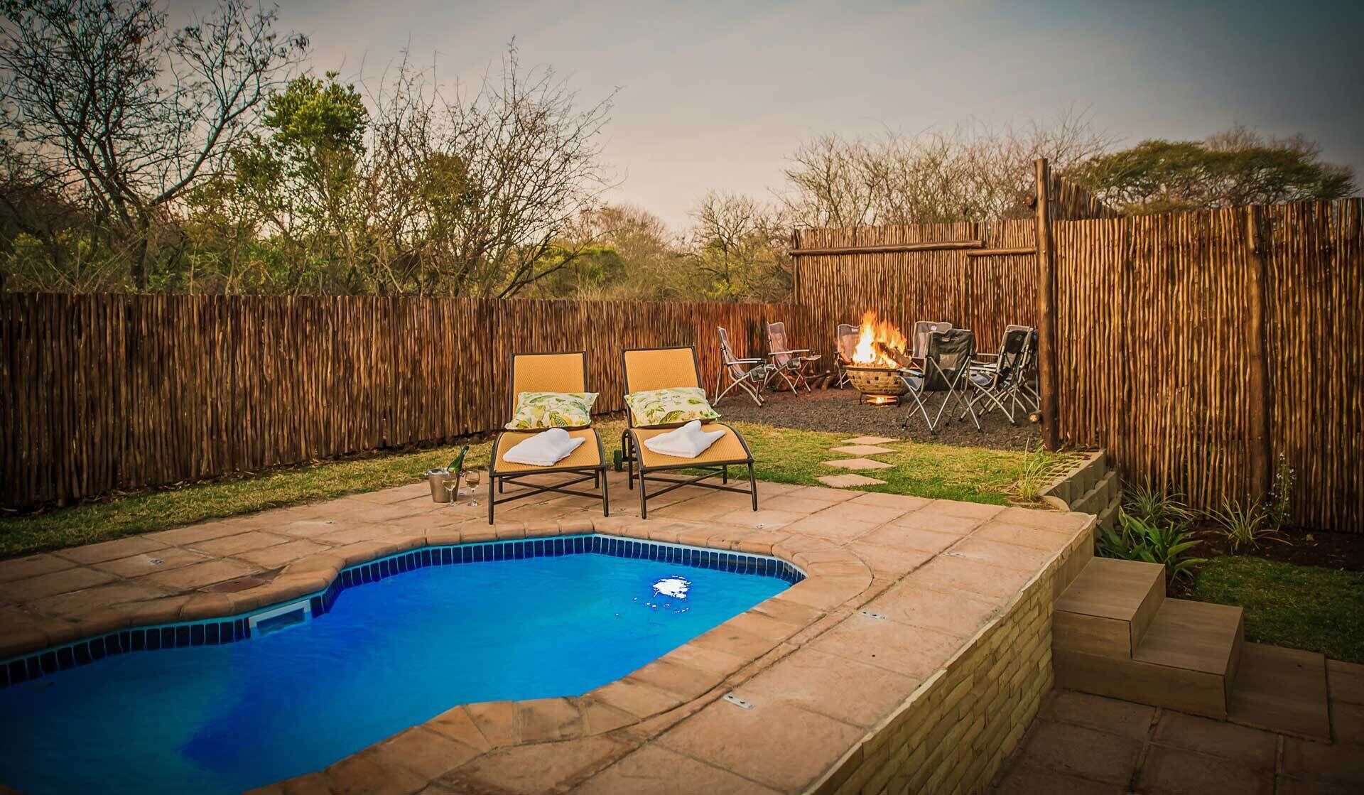 A backyard with a swimming pool, two lounge chairs, a fire pit, and a wooden fence.