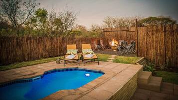 A backyard with a swimming pool, two lounge chairs, a fire pit, and a wooden fence.