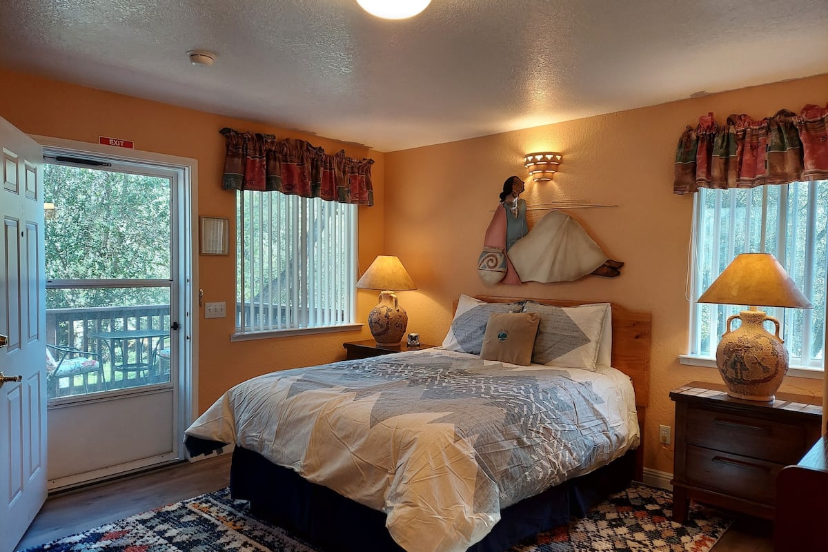 A bedroom with a large bed, two bedside lamps, a nightstand, a window with curtains, and a view of the outdoors.