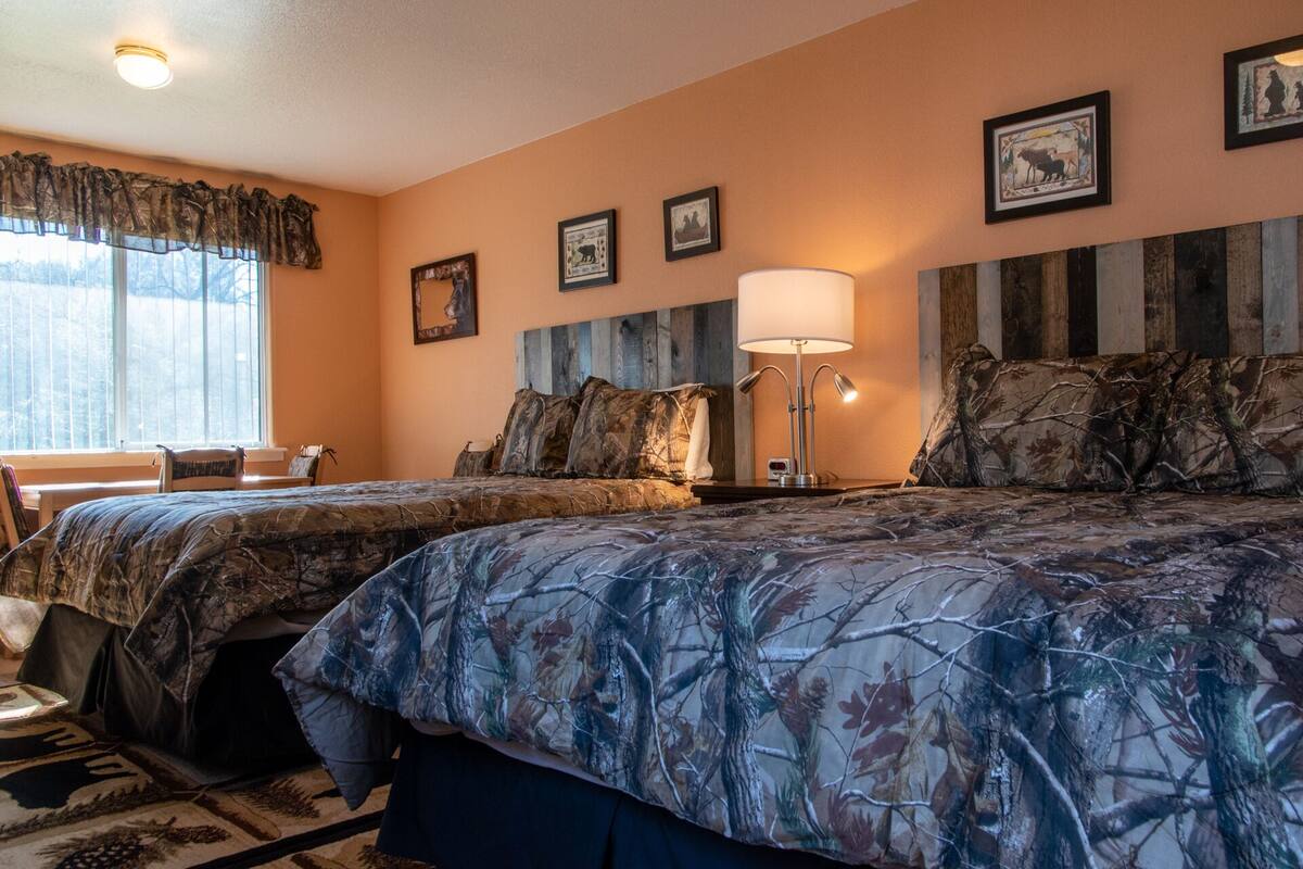 A room with two beds, a nightstand with a lamp, a window with curtains, and framed pictures on the wall.