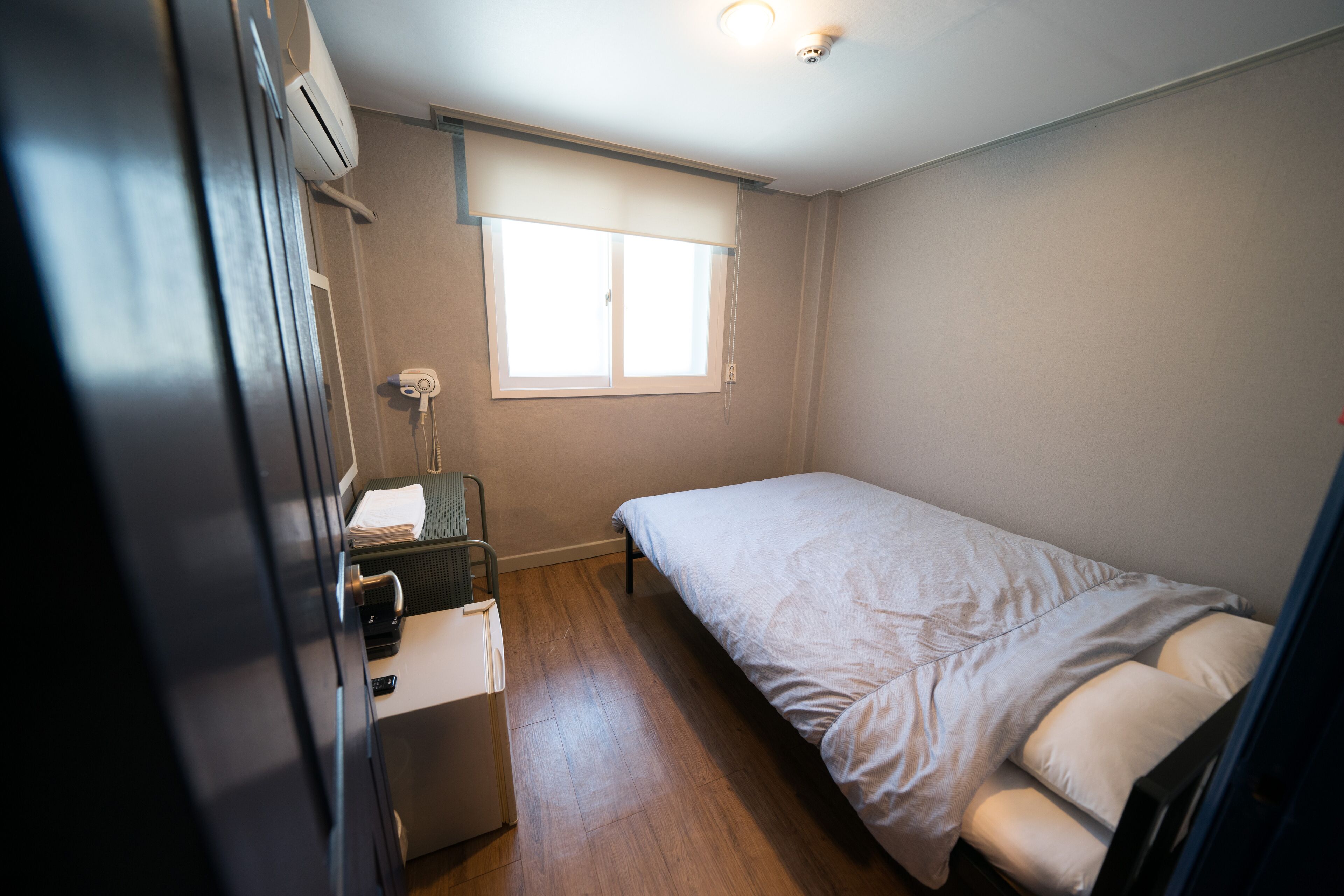 A bedroom with a bed, a window, a desk with a chair, and a small refrigerator.