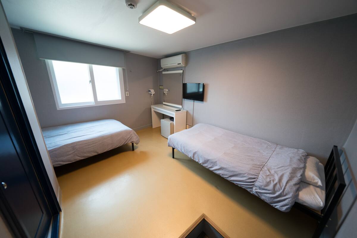 A small, well-lit room with two single beds, a desk, a flat-screen TV, and a window providing natural light.