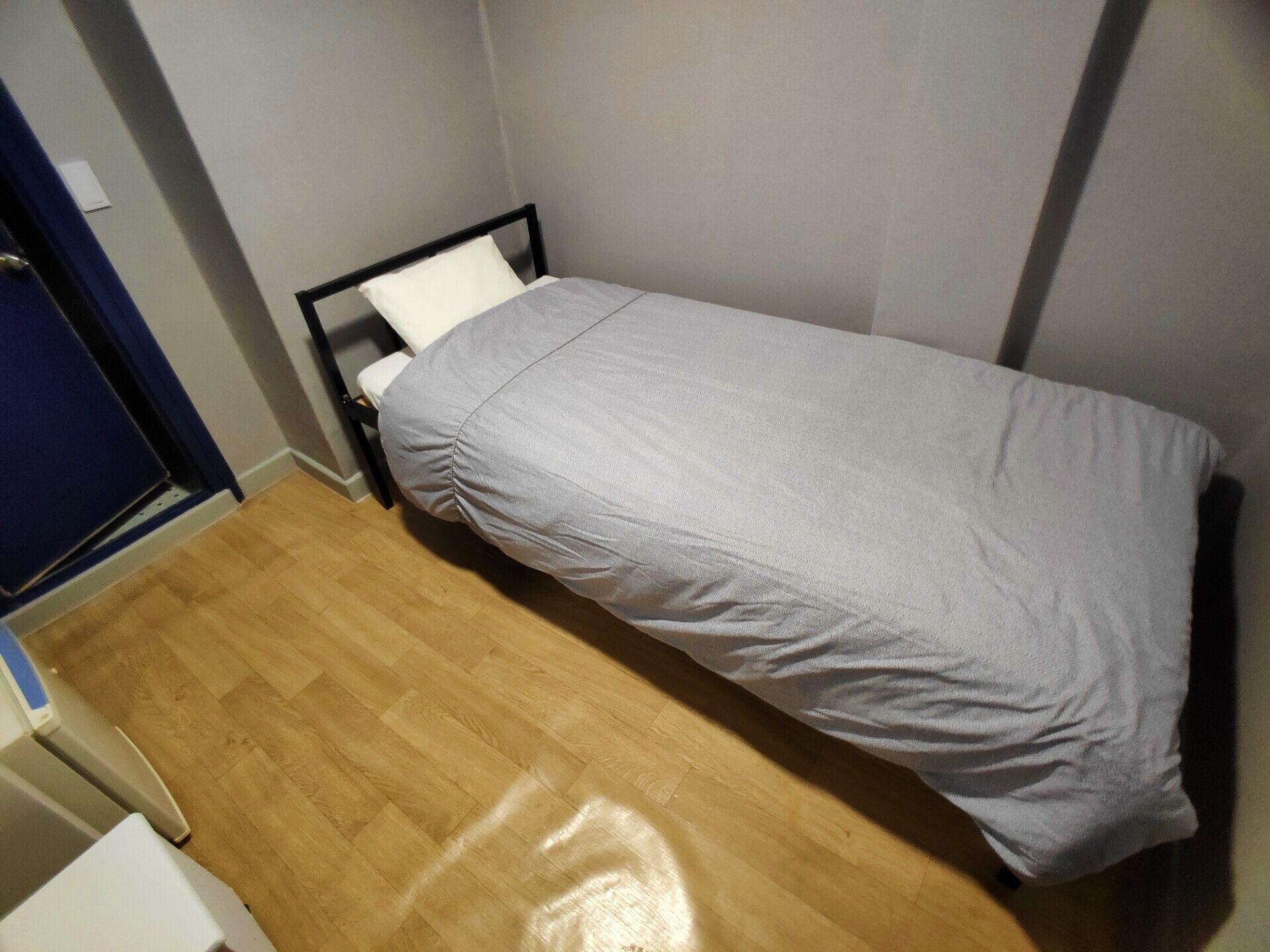 Una habitación pequeña con una cama individual, suelo de madera, pared blanca y una puerta con cortina azul.