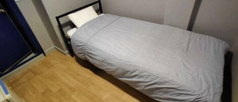 Una habitación pequeña con una cama individual, suelo de madera, pared blanca y una puerta con cortina azul.