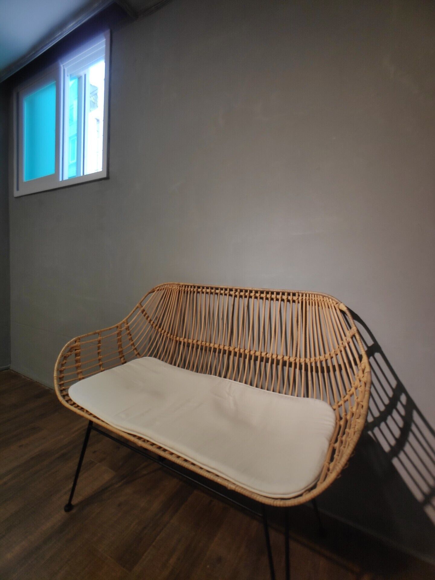 A wicker chair with a white cushion in a room with wooden flooring and a window.