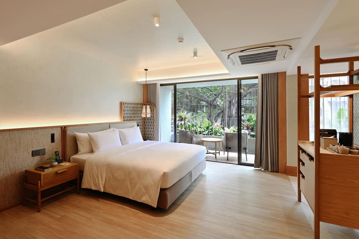 A modern hotel room with a large bed, a bedside table, a desk, and a balcony with a view of greenery.