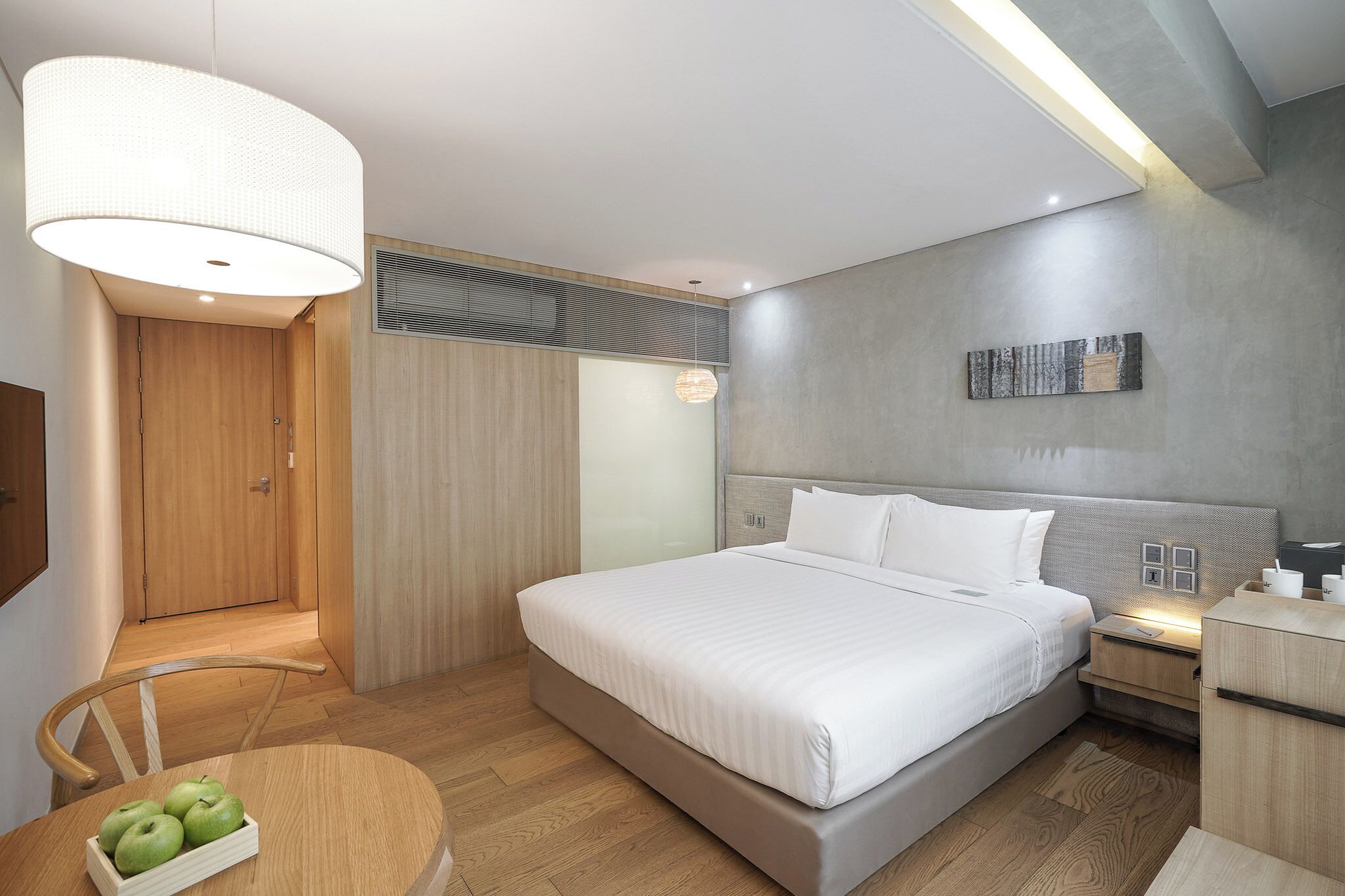A modern hotel room with a large bed, bedside tables, a round dining table with apples, and a large hanging light fixture.
