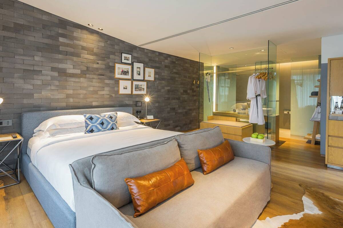 A modern bedroom with a large bed, a sofa, a glass-enclosed bathroom, and a brick-patterned wall.