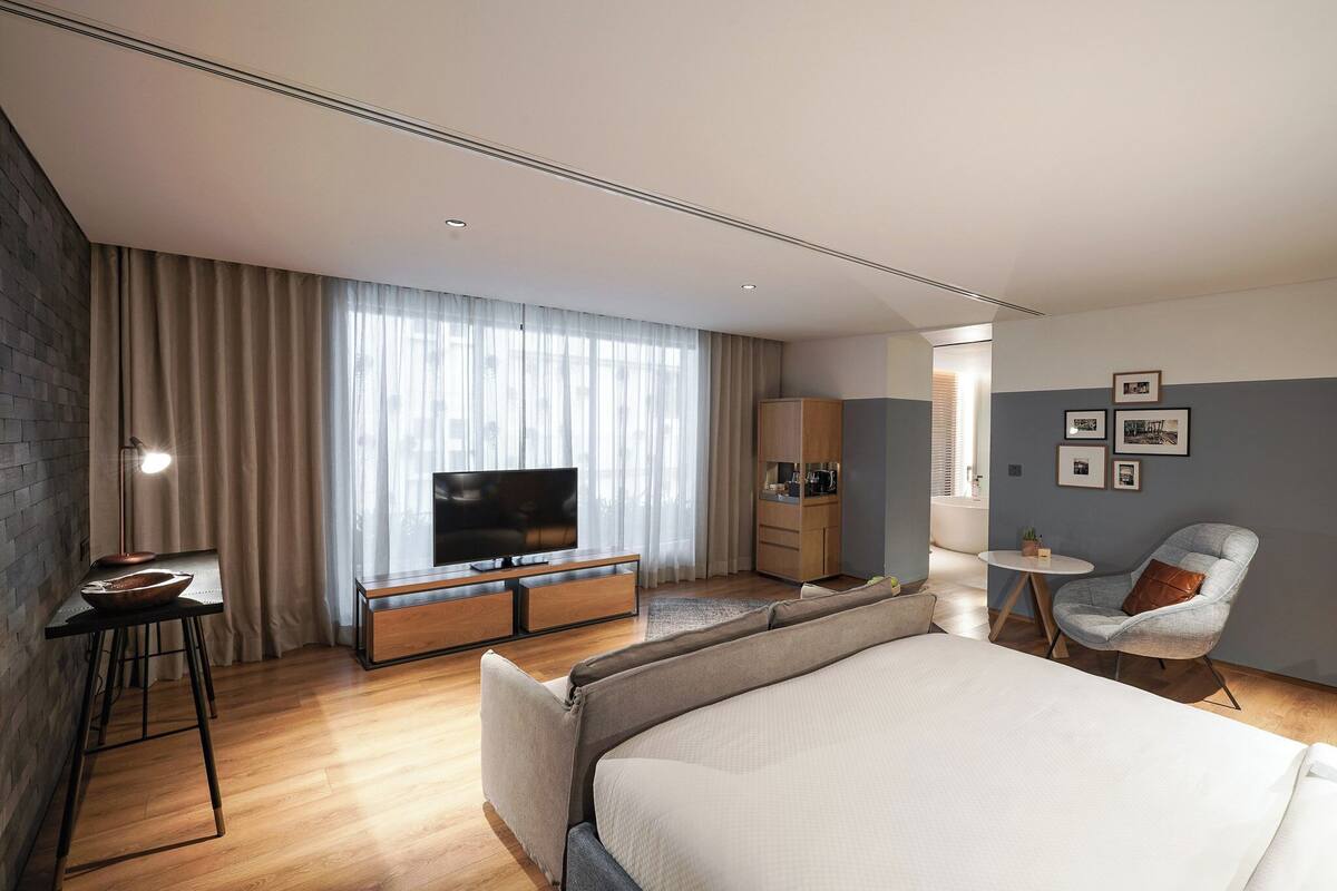 A modern hotel room with a large bed, a TV on a wooden console, a small table with a bowl, a chair, and a round table with a lamp.