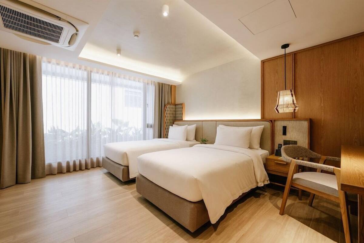 A hotel room with two beds, a wooden headboard, a bedside table, a chair, and a large window with curtains.