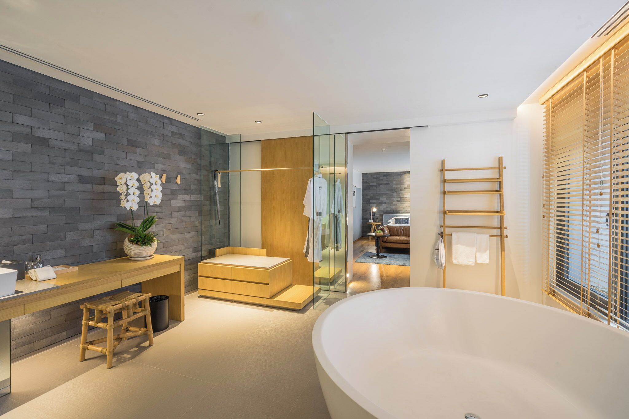 A modern bathroom with a large bathtub, a wooden vanity, a glass shower enclosure, and a view into the living area.