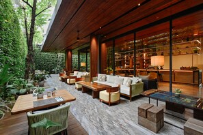 A modern outdoor lounge area with wooden furniture, a glass-enclosed bar, and lush greenery. - Ad Lib Hotel Bangkok (Bangkok)
