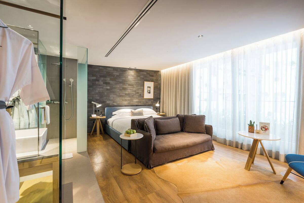 A modern hotel room with a glass-enclosed shower, a bed with a gray headboard, a brown sofa, a small round table, and a large window with sheer curtains.