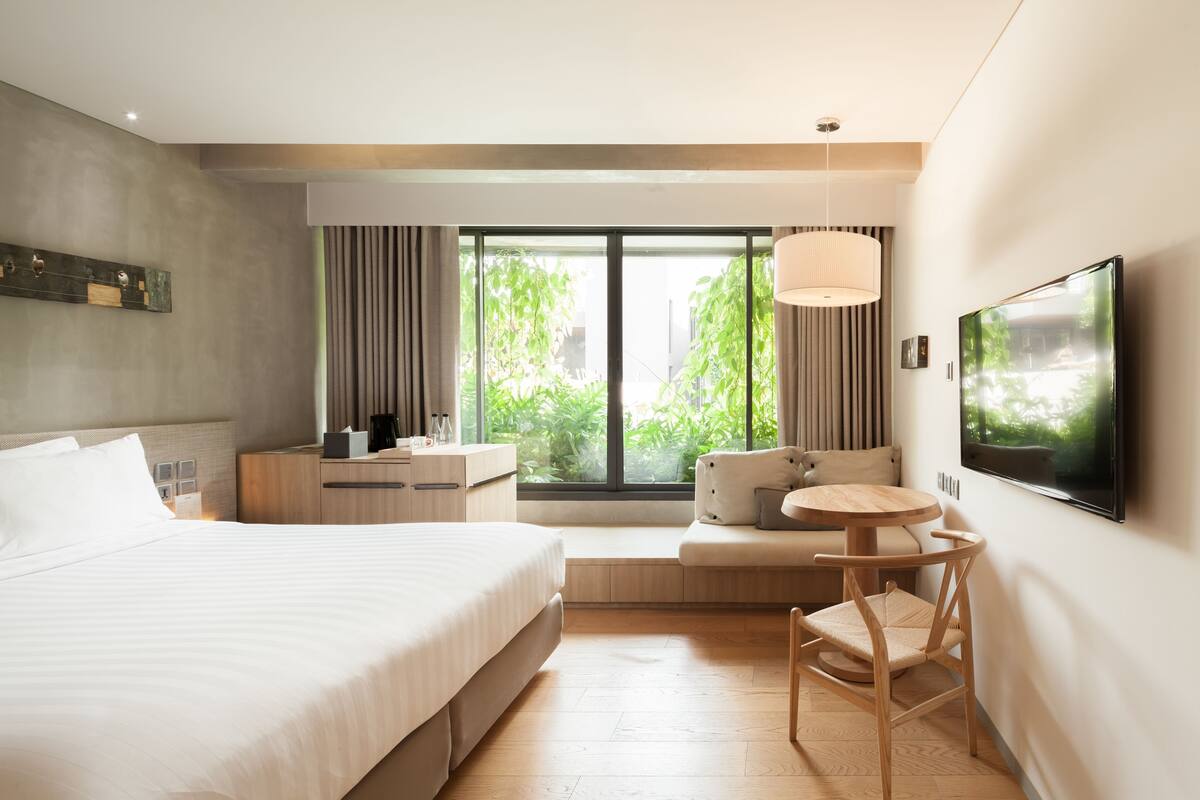 A modern hotel room with a large bed, a seating area with a table, a TV mounted on the wall, and a window with a view of greenery.