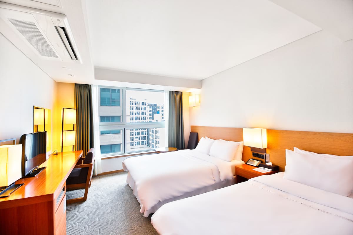 A hotel room with two beds, a desk, a computer, and a window with a view of buildings.