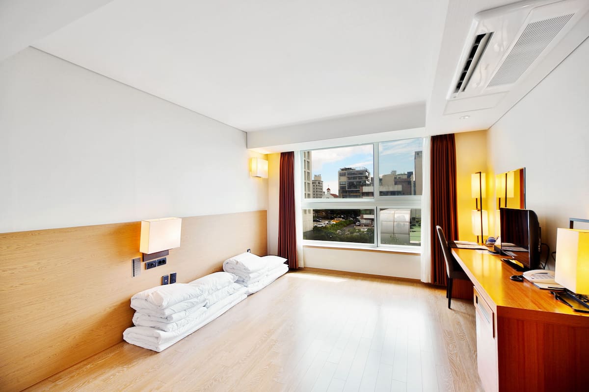 A hotel room with a wooden headboard, a desk with a computer, a TV, and a large window with curtains.