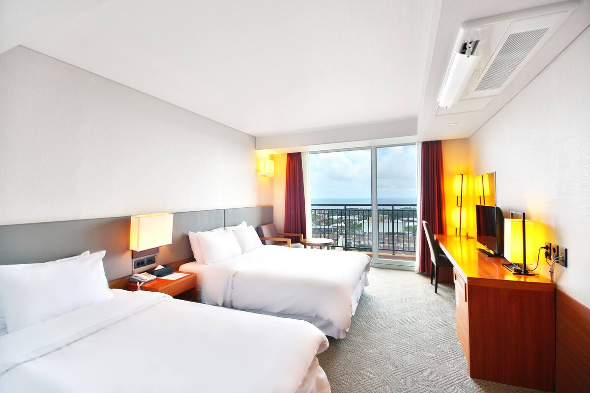 A hotel room with two beds, a desk, a TV, and a balcony with a view.