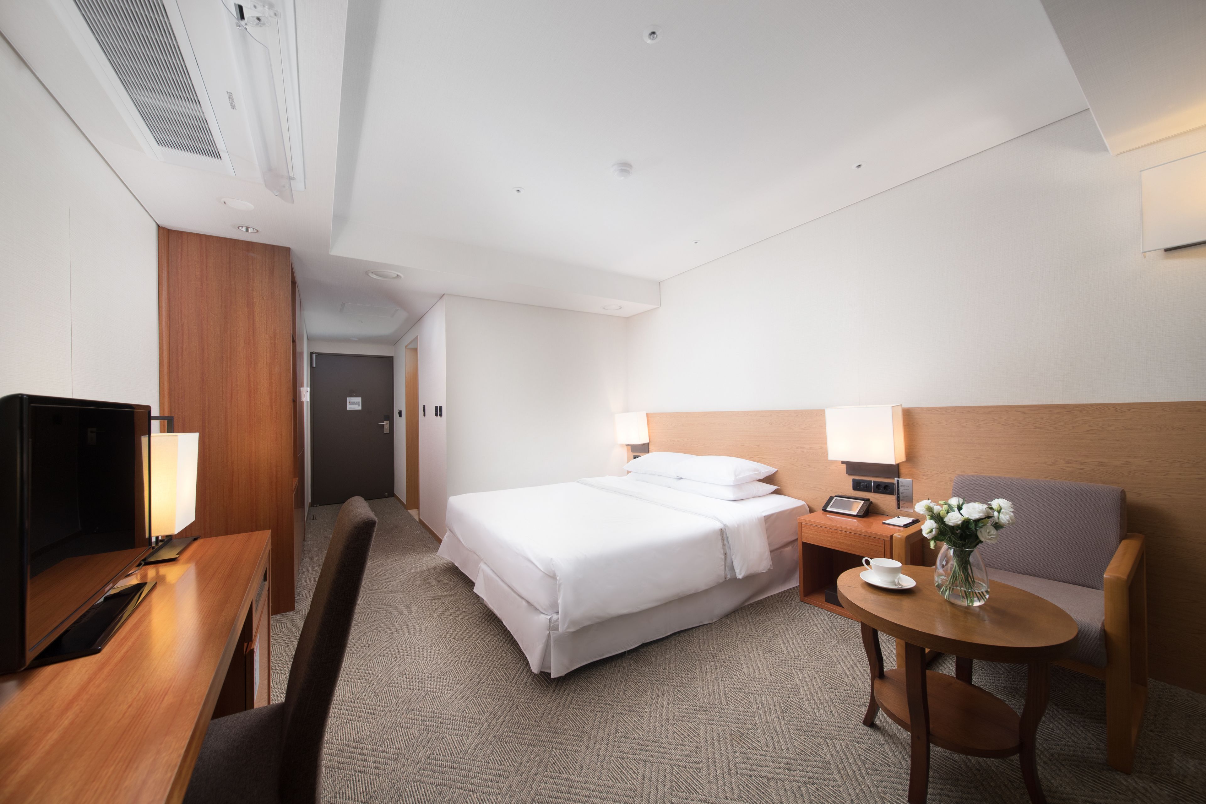 A hotel room with a bed, a desk with a lamp, a chair, a small table with a vase, and a television.