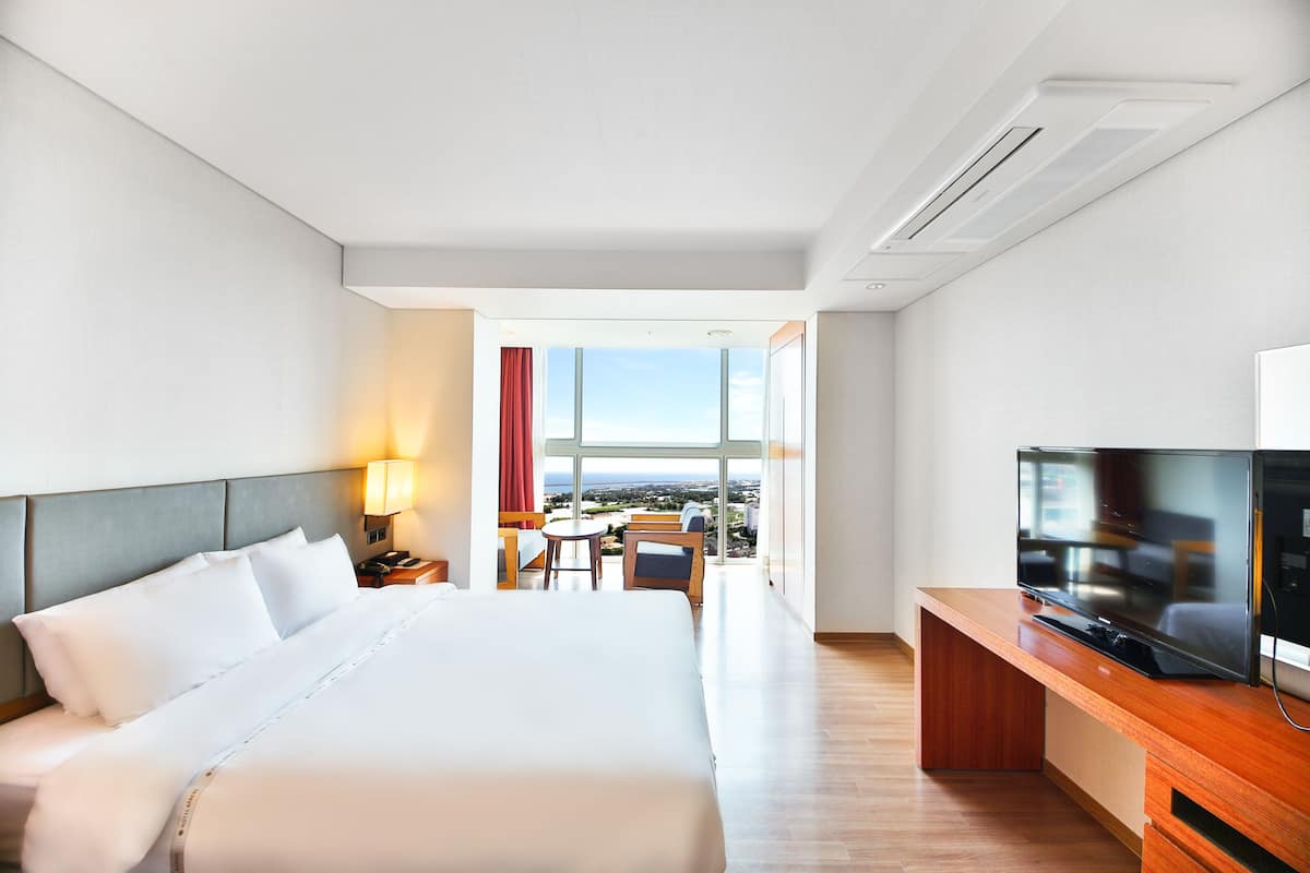 A hotel room with a large bed, a desk with a TV, a small table with chairs, and a view of the city through large windows.