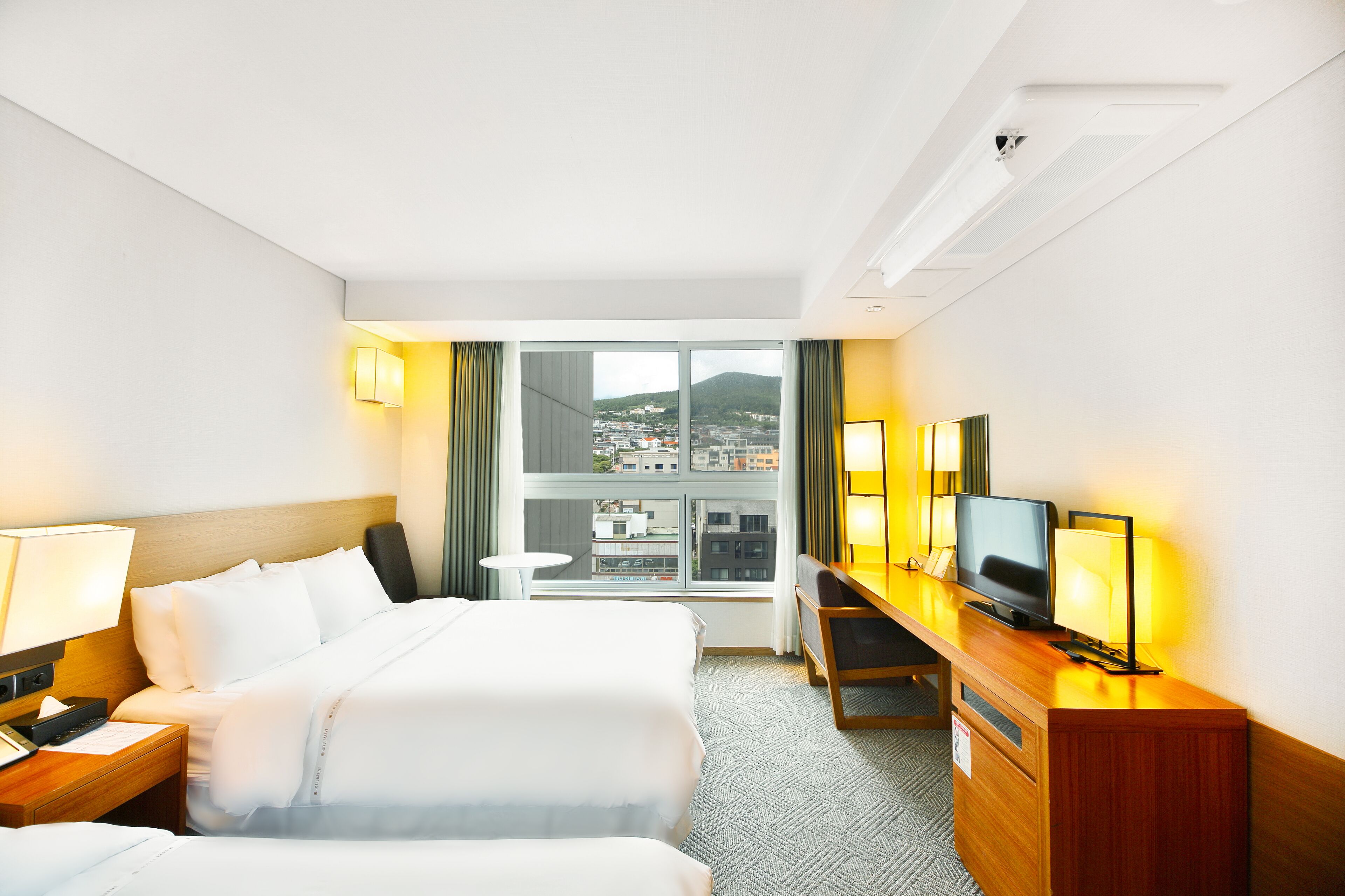 A hotel room with two beds, a desk with a TV, and a view of a cityscape through large windows.